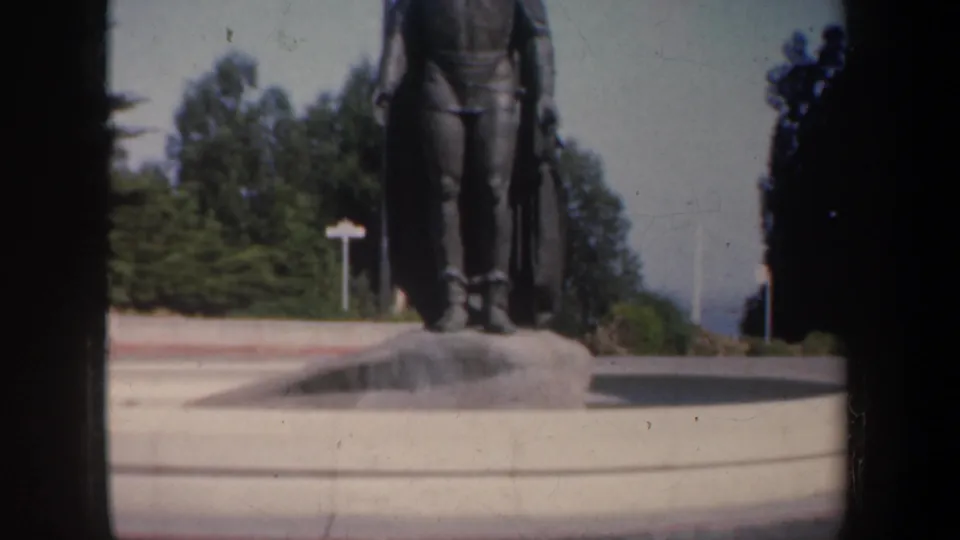 San Francisco home-movie frame with a bronze statue on a pedestal, surrounding trees, open paving, and a glimpse of blue water beyond.