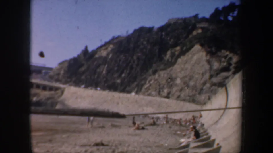 San Francisco coastal frame with a rocky cliff, curved seawall or road edge, beach sand, and distant people near the shore.