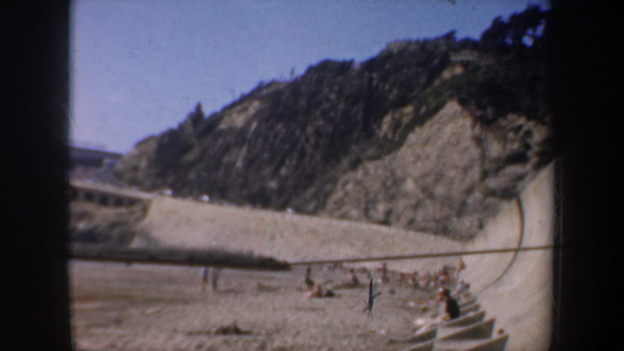 Sandy beach below a rocky cliff with a concrete wall and seated beachgoers near the shore.