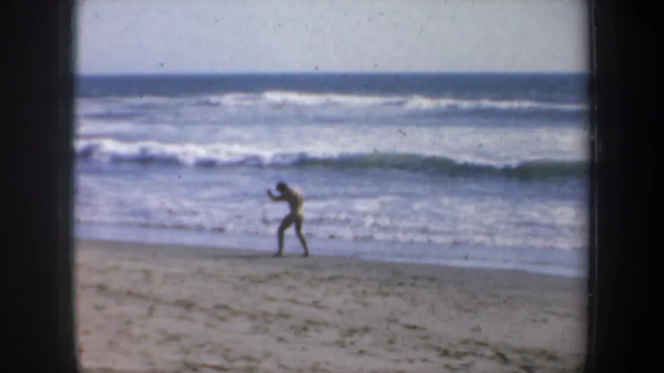 A man appears to shadowbox on a San Francisco beach in 1957, standing near the waterline as waves break behind him.