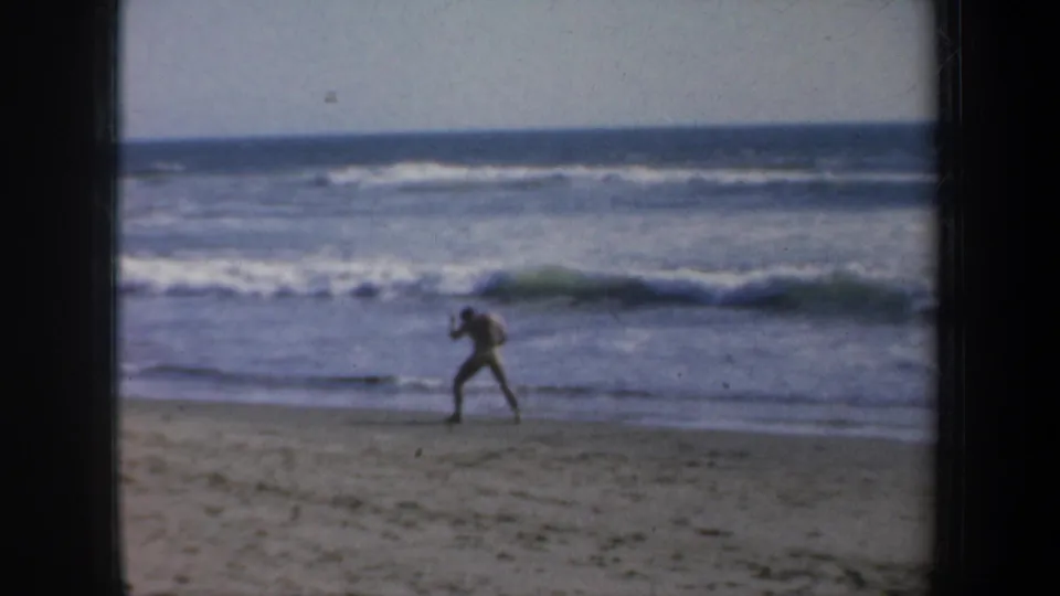 The adult figure continues shadowboxing on the 1957 San Francisco beach, filmed at a distance against low waves and open sand.