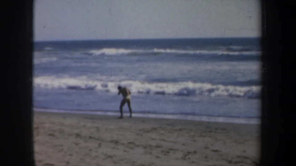 A distant adult man practices punches on a San Francisco beach in 1957, silhouetted against surf and a bright ocean horizon.