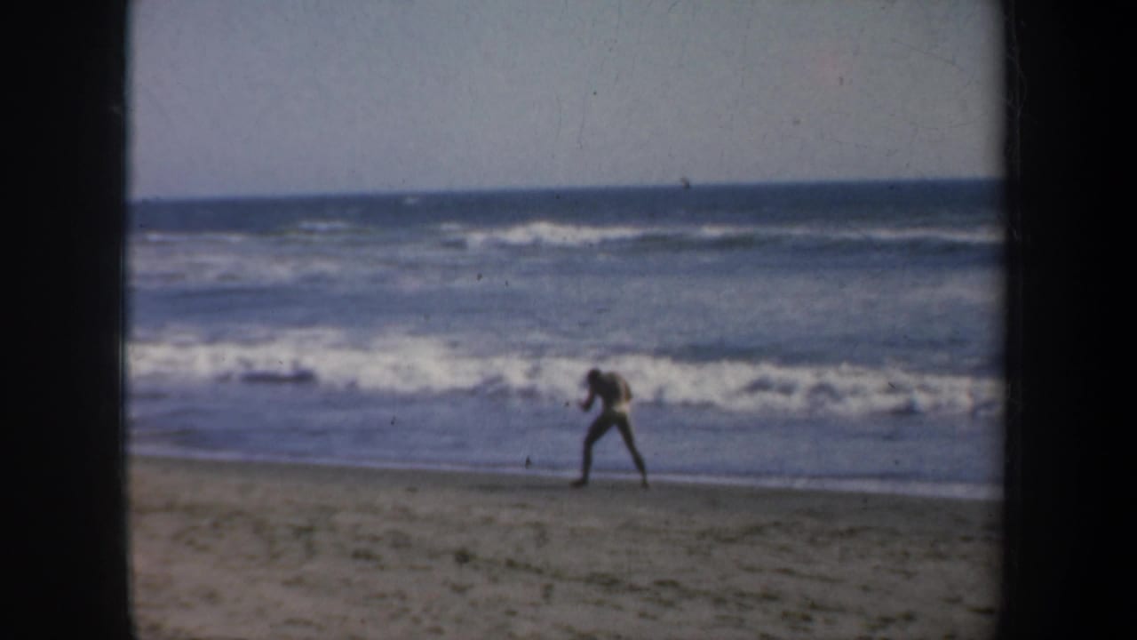 1957 San Francisco 8mm frame showing a distant figure near the surf on a broad beach with waves and dark film borders.