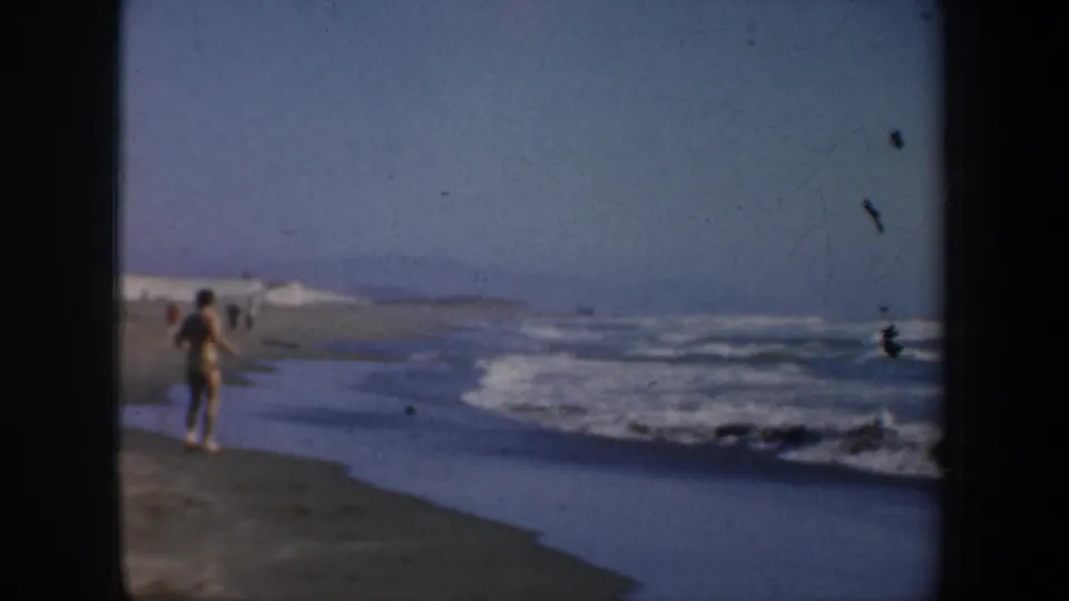 Home-movie view of a beach figure moving along the San Francisco shoreline, with surf rolling in and hazy beach in the distance.