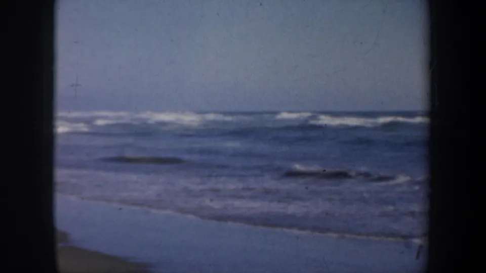 1957 San Francisco beach footage frame with rolling waves, reflective wet sand, pale sky, and visible 8mm gate edges.