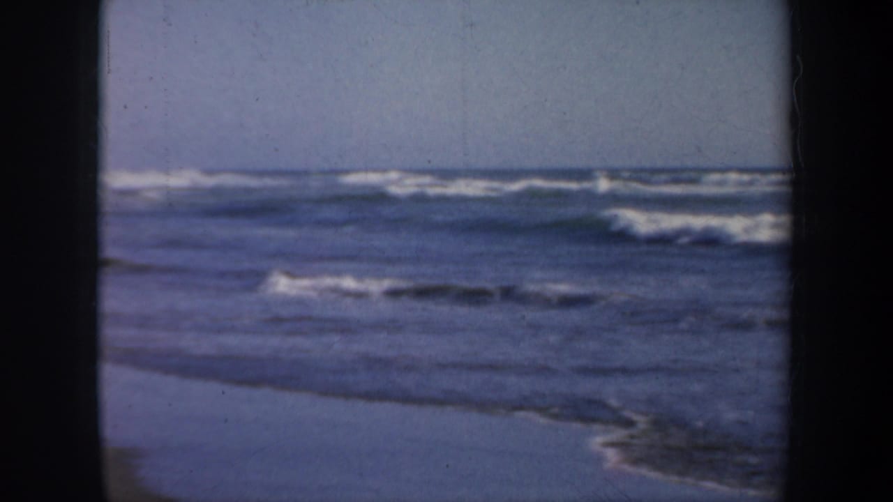 Ocean waves roll toward wet sand under a pale sky, framed by dark 8mm borders.