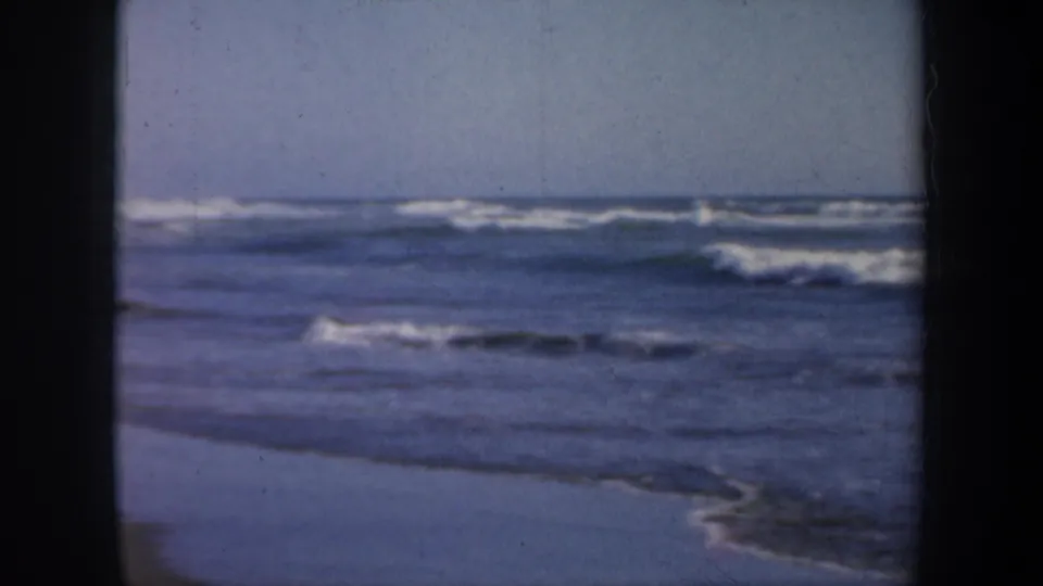 1957 San Francisco coastal home-movie frame showing low ocean surf, white foam, wet beach, and dark 8mm projector-mask edges.