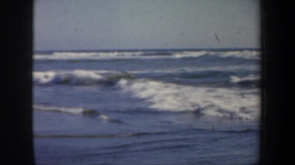 Mid-century 8mm frame from San Francisco showing white surf washing over wet beach beneath a pale coastal sky.