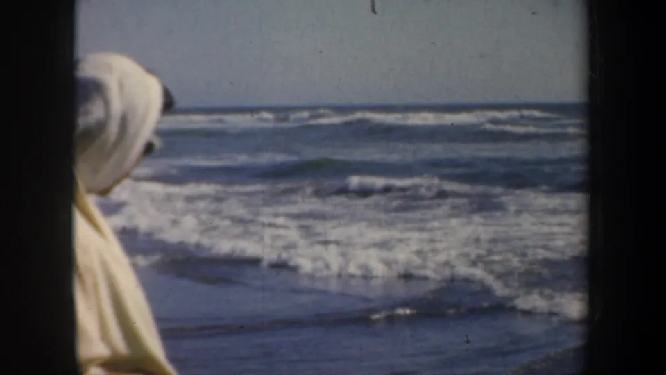 1957 San Francisco beach frame with a partly visible scarfed person at the edge of the shot and ocean surf in the background.