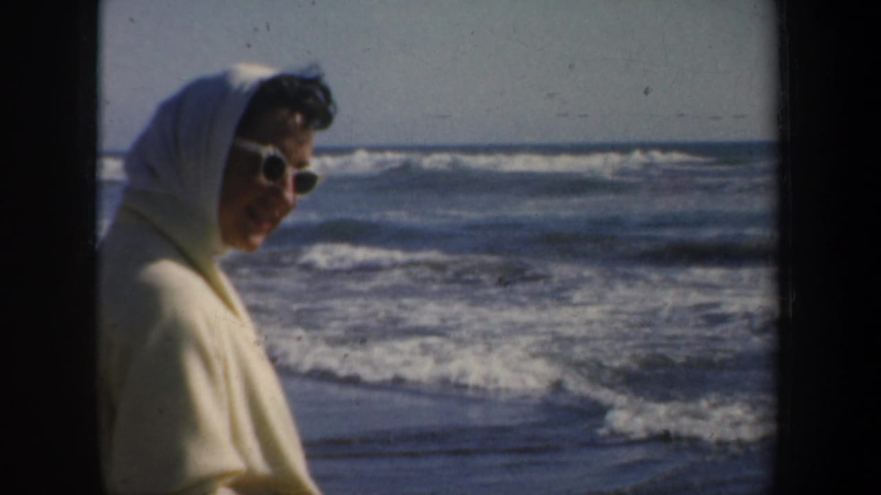 Woman in sunglasses and a light headscarf stands near ocean waves.