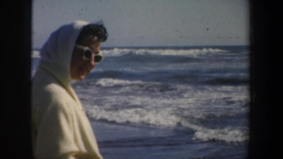 In 1957 San Francisco home-movie footage, a woman in sunglasses and a light headscarf looks toward breaking ocean waves.