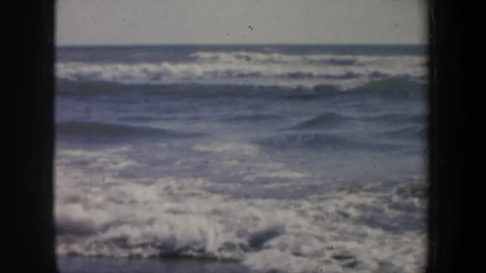 A 1957 San Francisco beach view shows low ocean waves and white foam moving toward shore in soft 8mm color.