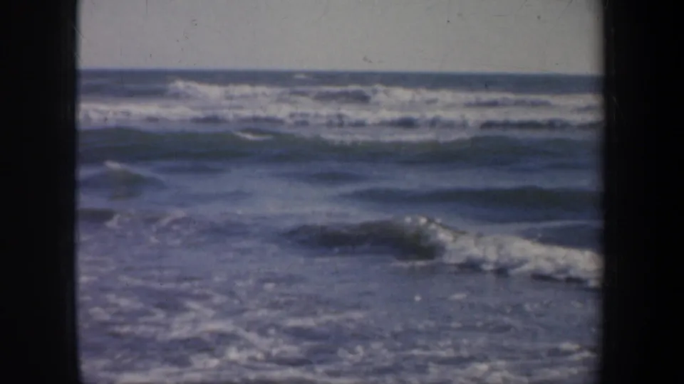 1957 San Francisco coastal home-movie frame showing surf beyond a pier railing, with dark structural edges framing the view.