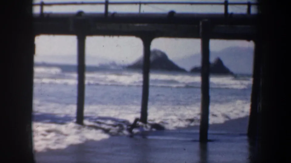 1957 San Francisco coastal frame showing pier posts over wet sand and surf, with distant rock forms visible through the structure.