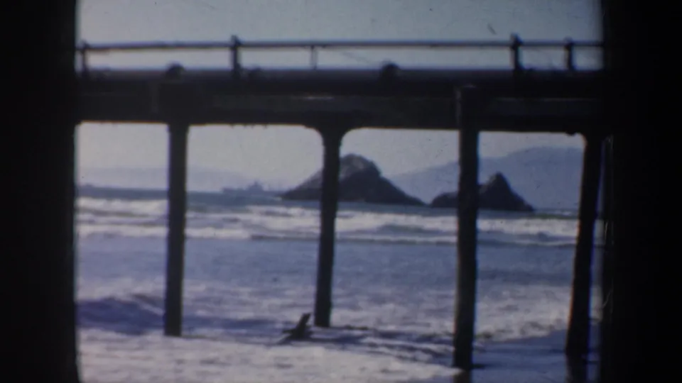 1957 San Francisco beach view from beneath or beside a pier, with posts, railing, surf, and distant rock formations.