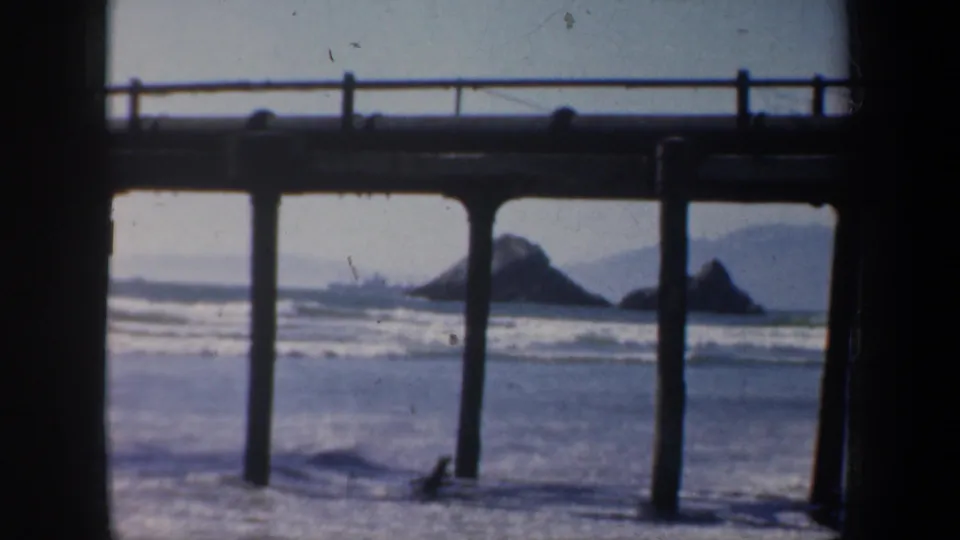 1957 San Francisco coastal view framed by silhouetted pier pilings, with waves and distant rocks beyond.