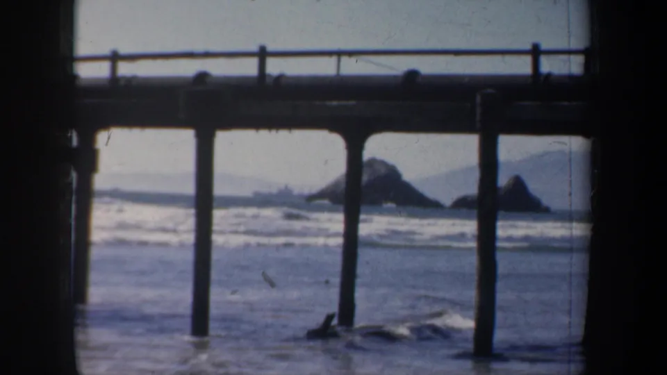 A mid-century 8mm coastal frame shows San Francisco surf beyond dark pier supports and rocky outcrops.