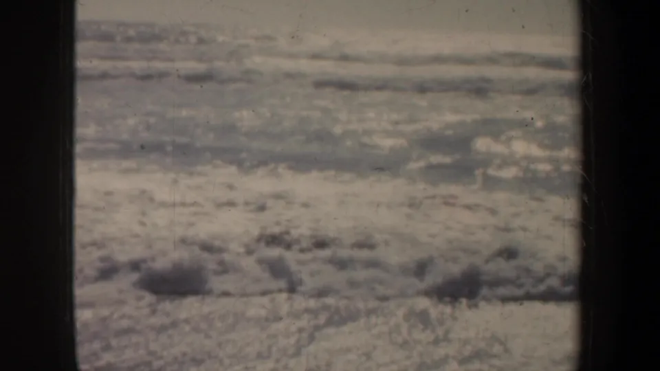 1957 coastal home-movie frame of surf washing across wet sand, seen from beneath a San Francisco pier.