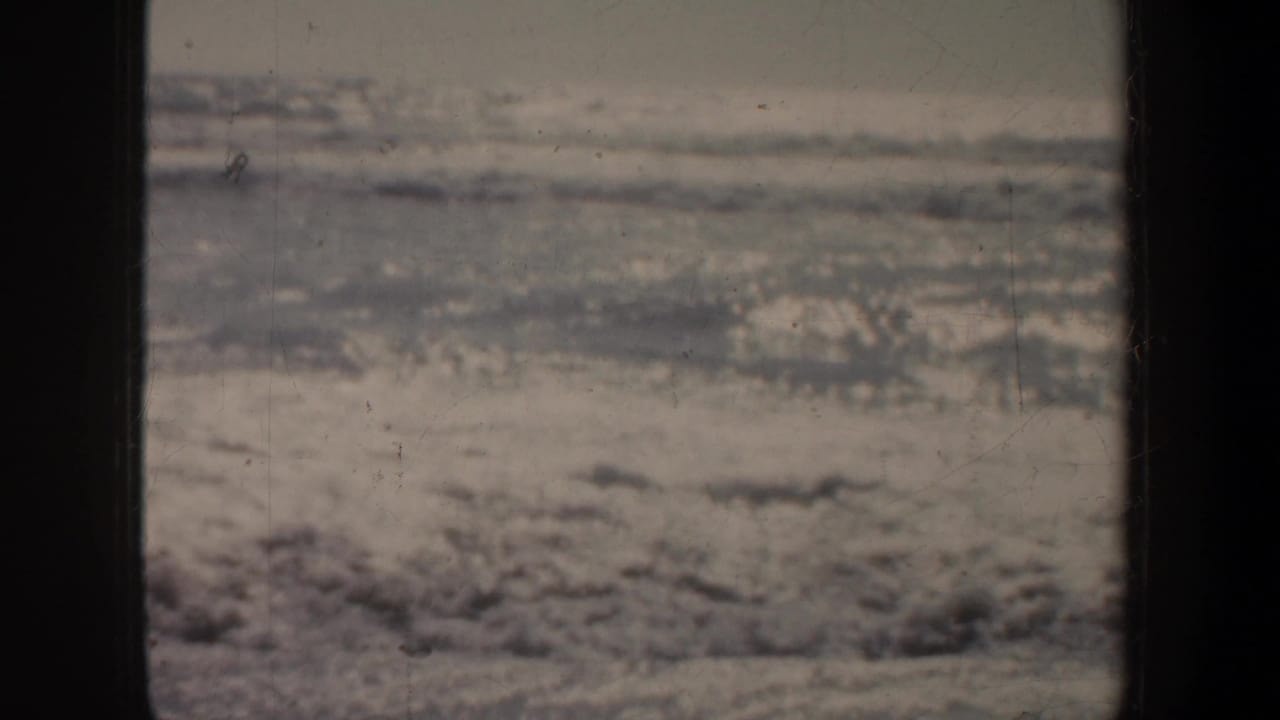 1957 San Francisco 8mm frame showing muted ocean water, thin foam, and wet sand seen from a fixed shoreline viewpoint.