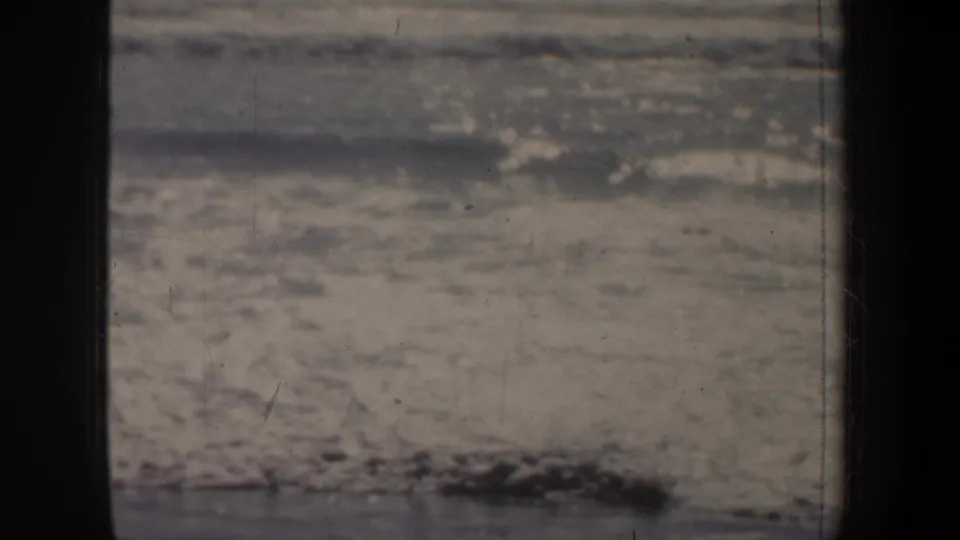 1957 San Francisco 8mm frame with rippled water, pale foam, and dark wet shoreline texture under soft exposure.