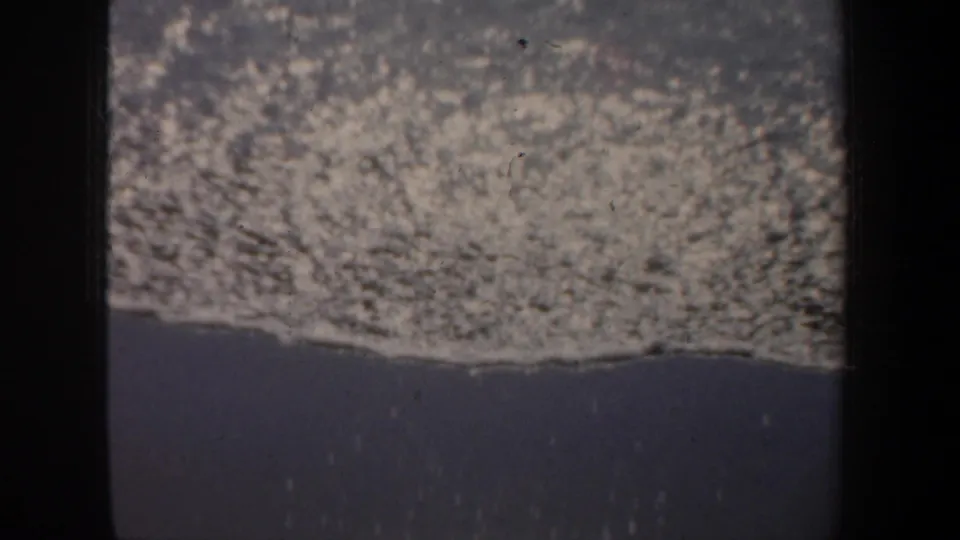 1957 San Francisco 8mm frame showing foamy surf washing over dark wet sand with visible grain and gate-edge shadow.