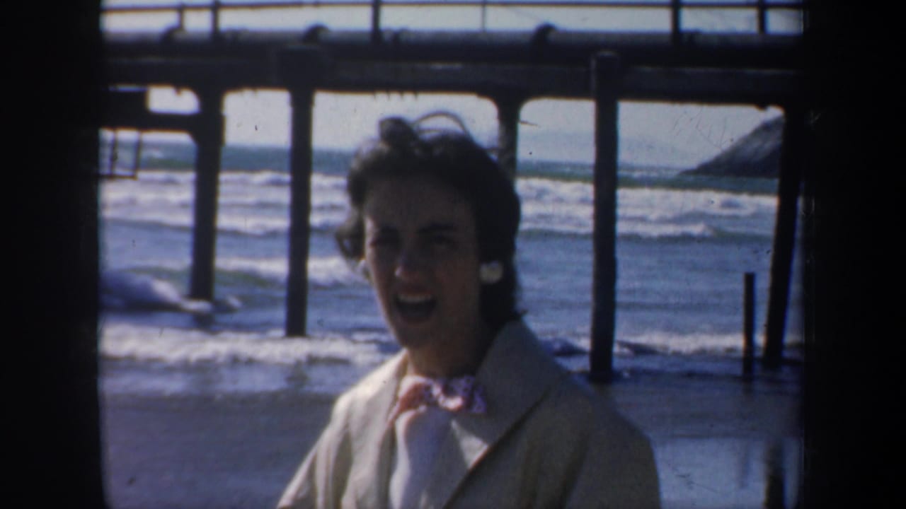 Woman in a light coat smiling under a pier with ocean waves behind her.