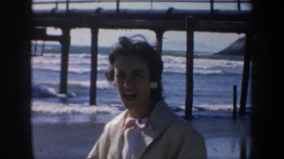 In 1957 San Francisco home-movie footage, a woman in a light coat stands under a wooden pier as surf rolls in behind her.