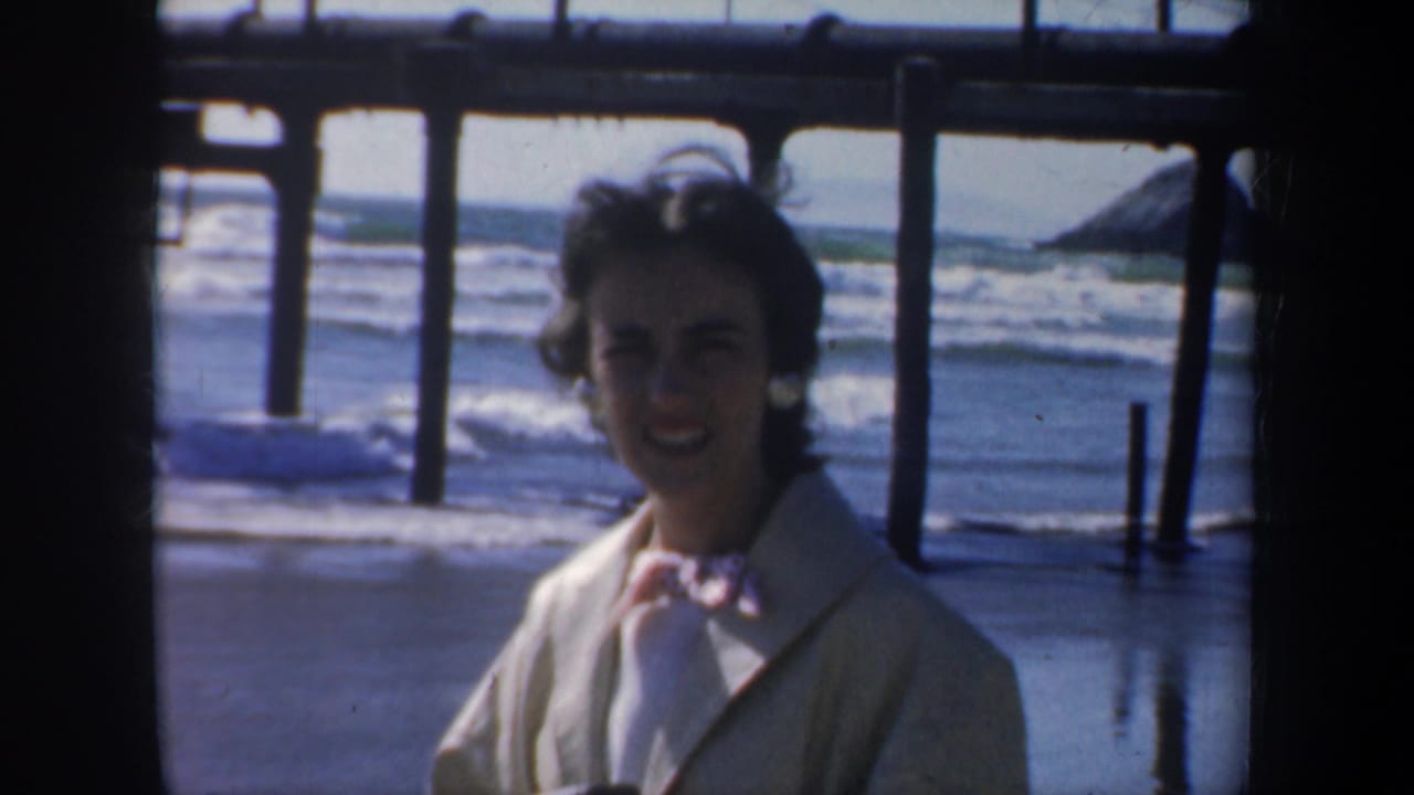 Woman facing the camera under a pier with waves and pilings behind her.
