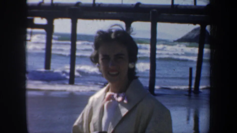 A woman in a light-colored coat faces the camera under a San Francisco pier, with ocean surf and wooden supports visible behind her.