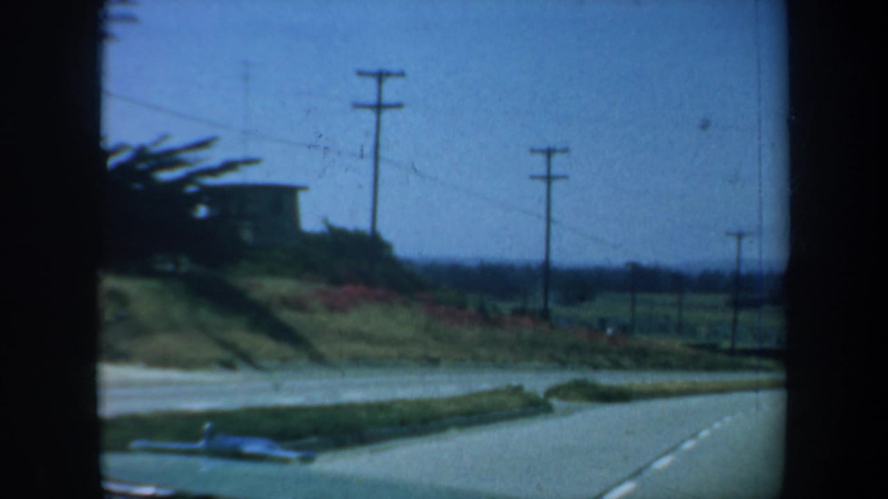 Road curving past a grassy hillside with utility poles and a small building.