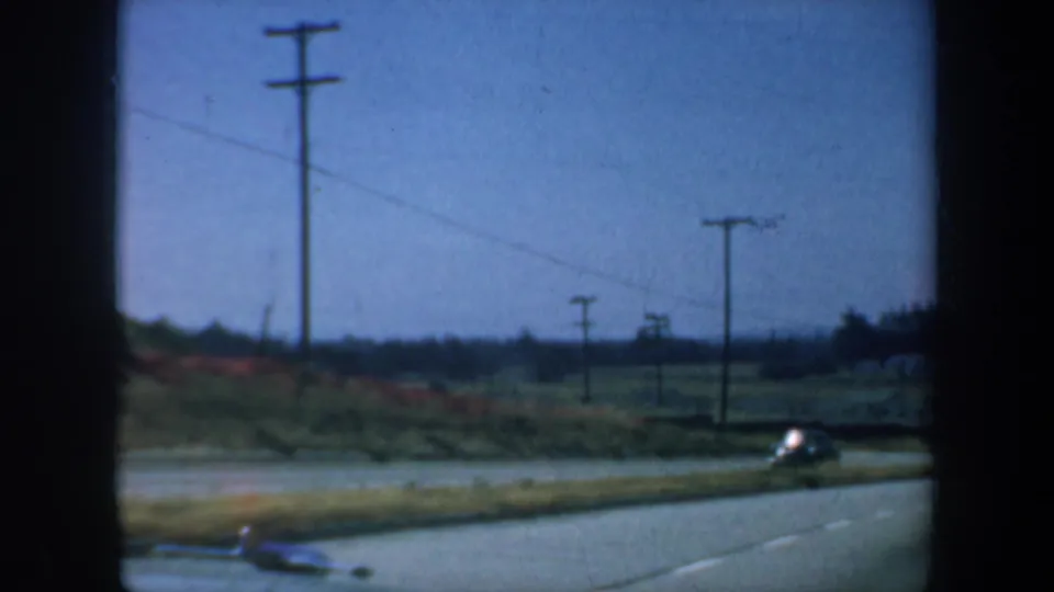 1957 home-movie roadside frame with utility poles, hillside vegetation, and a small car passing at the edge.