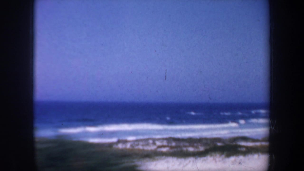 Blue ocean horizon and white surf beyond a sandy shoreline, partly framed by dark film edges.
