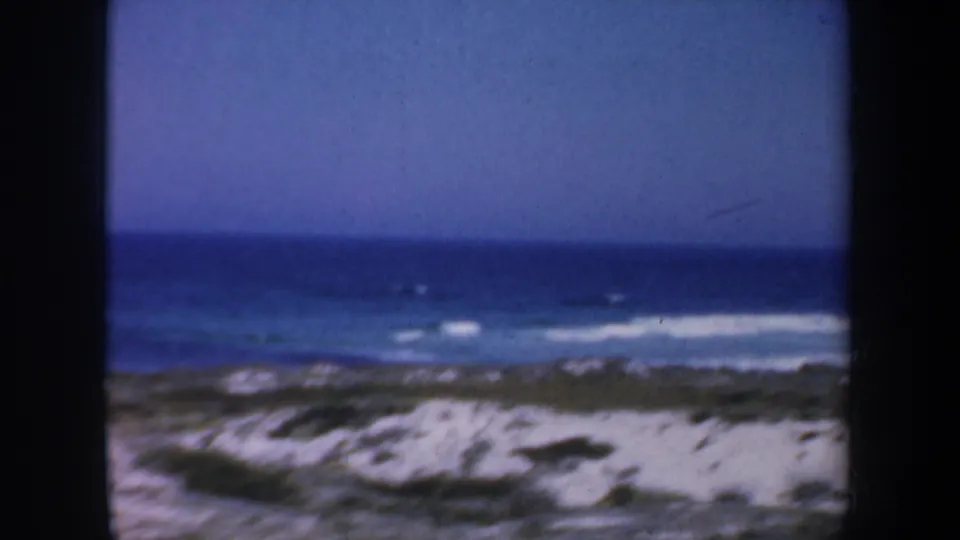 1957 8mm coastal frame showing blue water, white wave lines, pale sand, dune vegetation, and train-window-like borders.