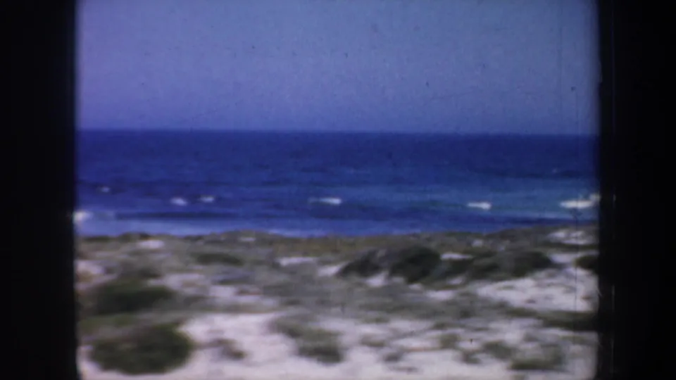 Soft 1957 8mm view of a California coastal landscape with dunes, scrub, blue ocean, and white surf lines in the distance.