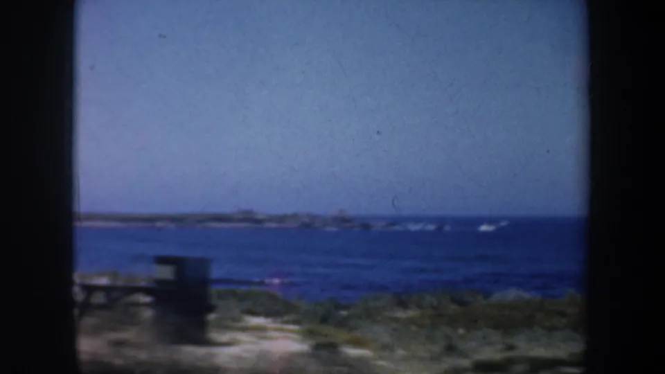 1957 8mm coastal frame showing blue water, distant shoreline, sandy ground, and a dark foreground edge.