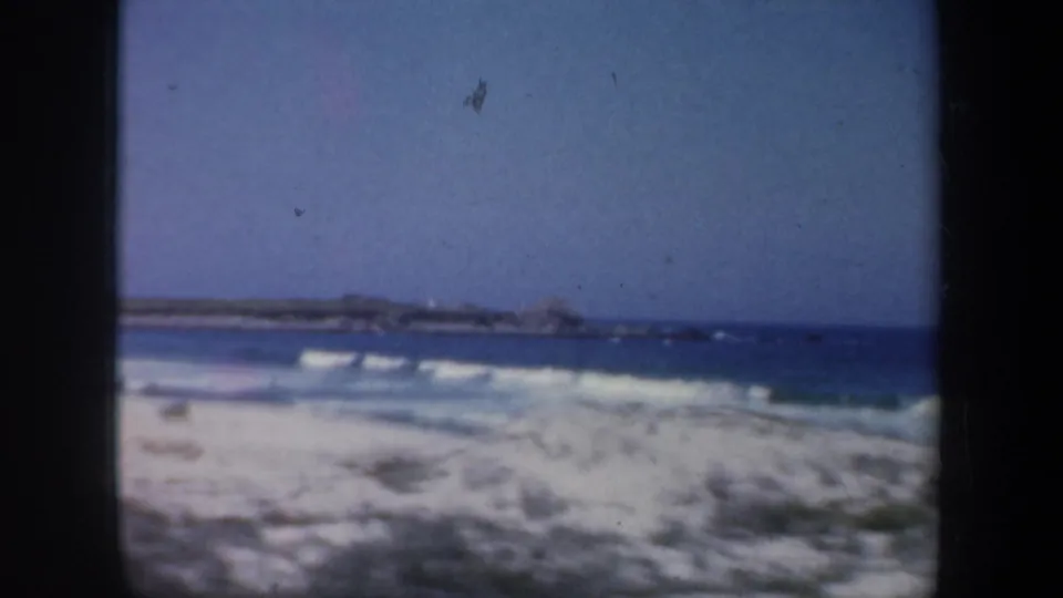 1957 San Francisco-area beach view with tan dunes, blue ocean, white surf, and a distant rocky shoreline.