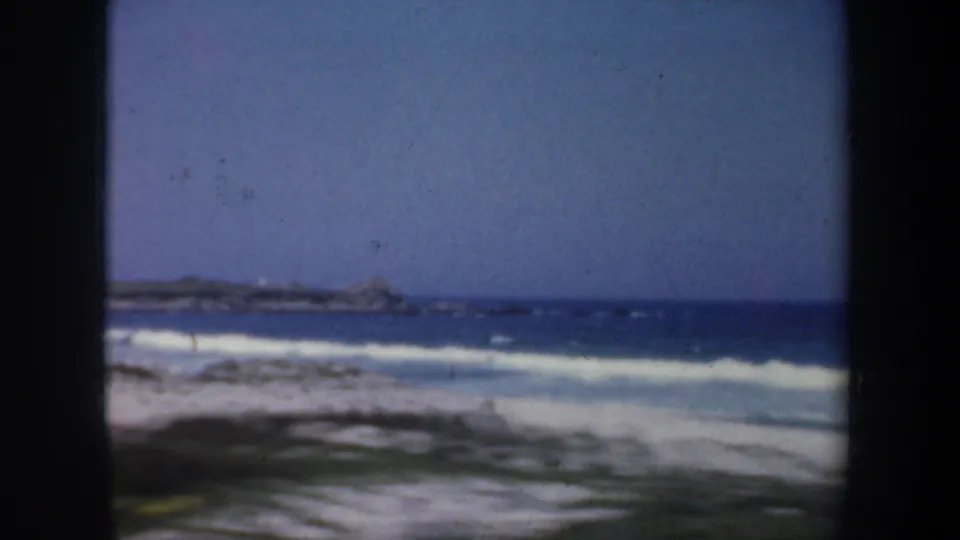 8mm frame of the San Francisco coast showing white surf, pale sand, blue water, and a low distant shoreline under clear sky.
