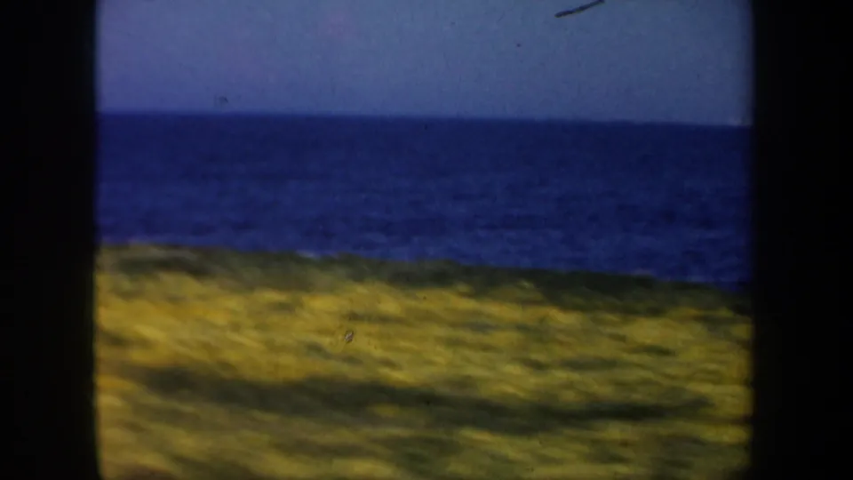 Moving 8mm view of grassy coastal dunes with deep blue ocean and clear sky, consistent with the 1957 San Francisco beach-drive metadata.