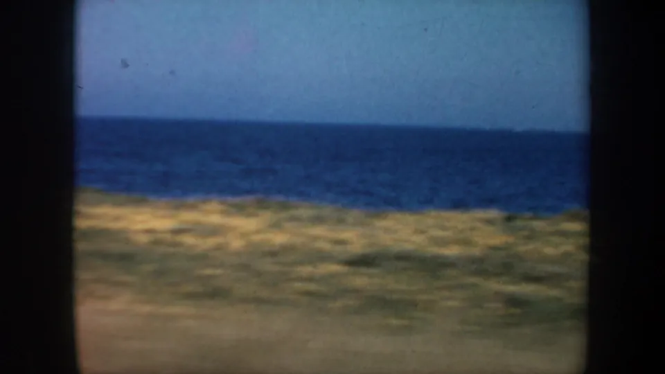 Coastal shoreline and open ocean pass by in a 1957 8mm San Francisco-area home-movie frame, with motion blur and dark window edges.
