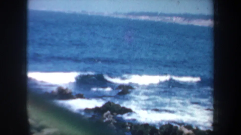 1957 8mm coastal frame showing waves breaking around dark rocks, blue water, and a distant shoreline in the San Francisco-area footage.