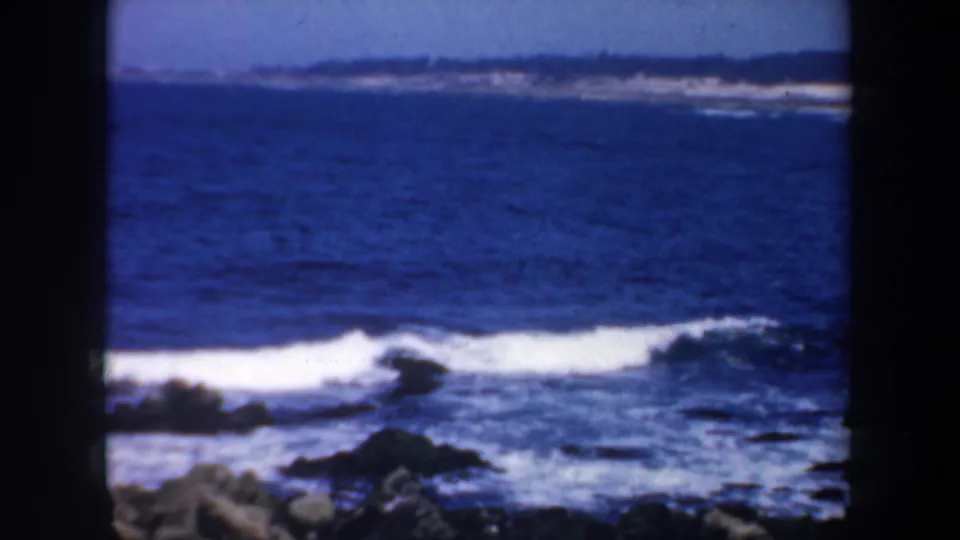 Rocky coastal surf in the 1957 San Francisco-area home movie, with white waves, dark rocks, blue water, and a distant shoreline.