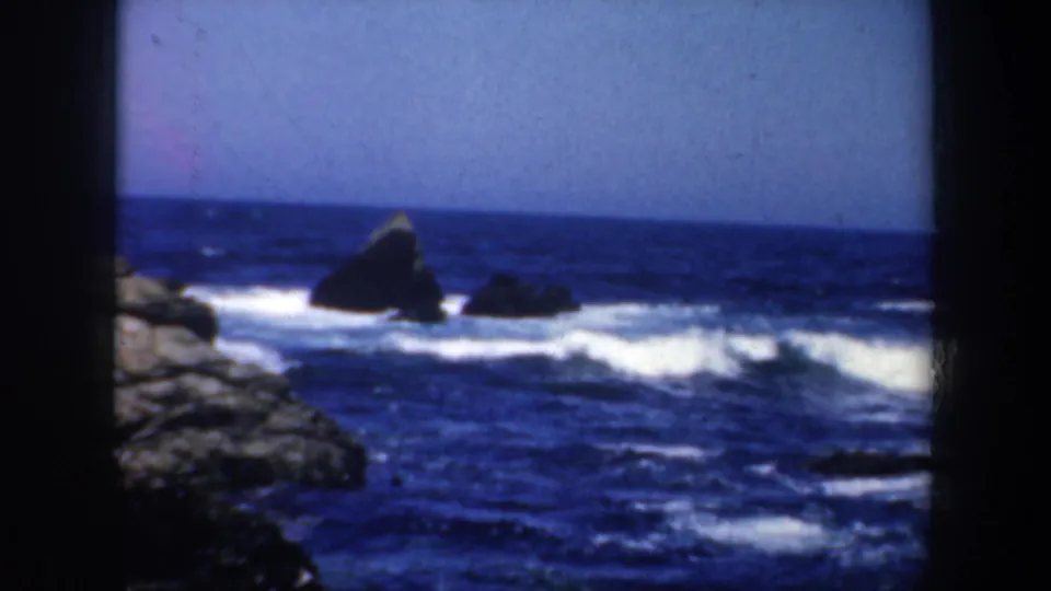 1957 San Francisco-area 8mm frame showing a pointed offshore rock, blue ocean, white surf, and rocky foreground.