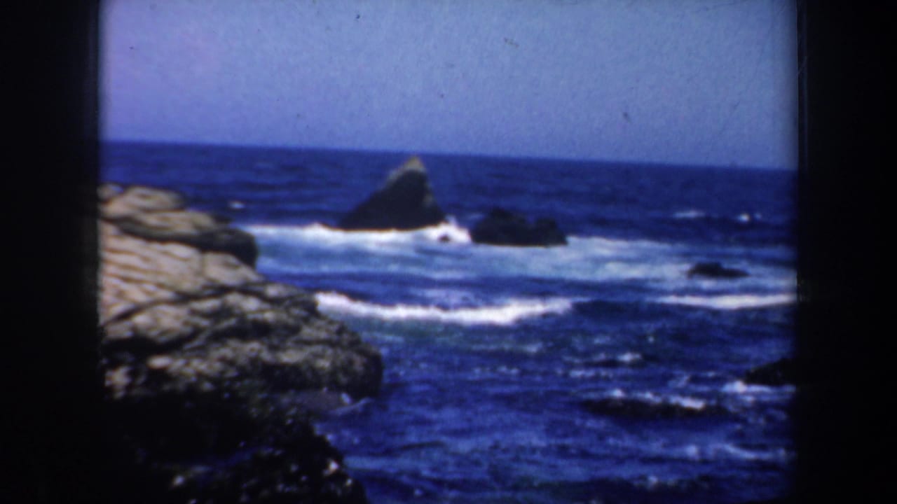 1957 8mm San Francisco-area coastal view with dark rocks, blue ocean, white foam, and a pale horizon.