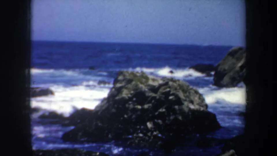 1957 8mm San Francisco coastal view centered on a large dark rock with foamy surf and blue water beyond.