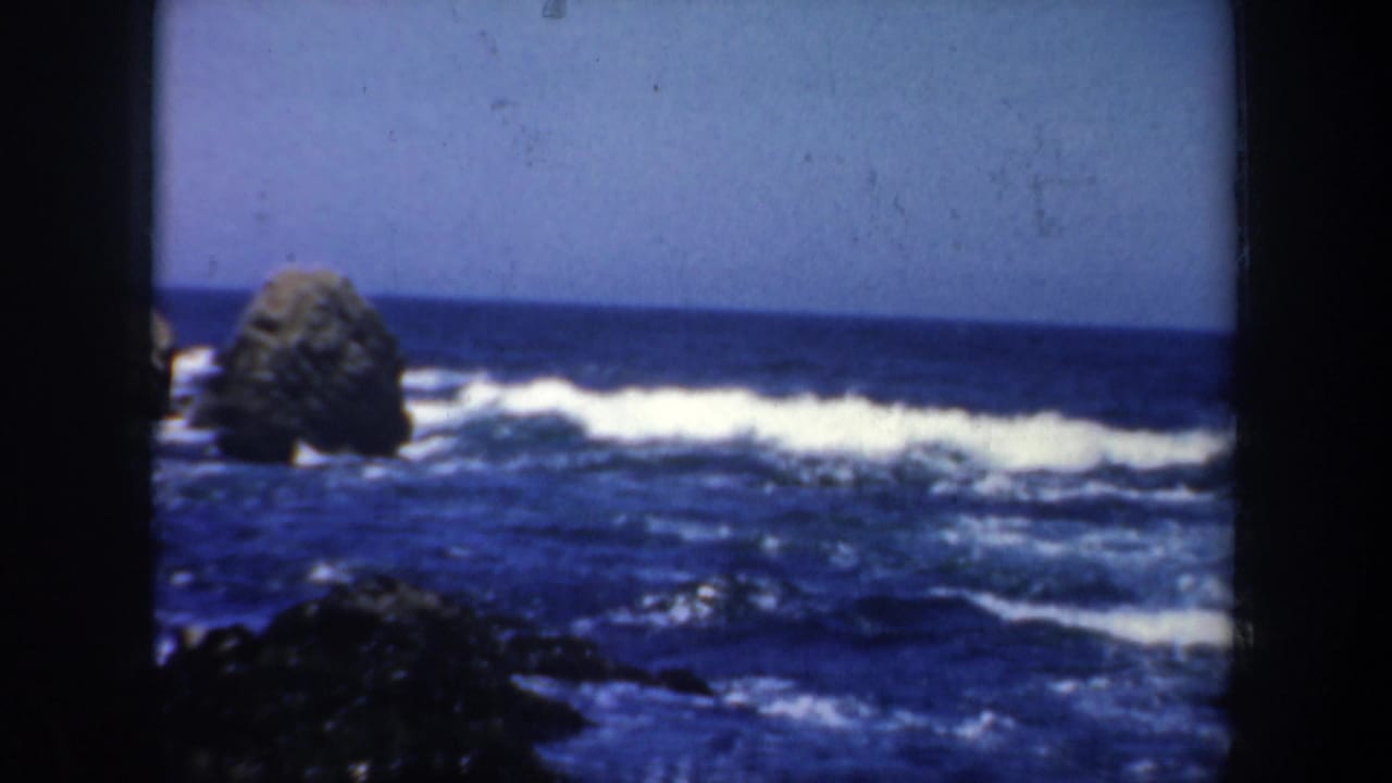 1957 San Francisco 8mm frame showing white surf breaking around a dark offshore rock along a rugged coastline.