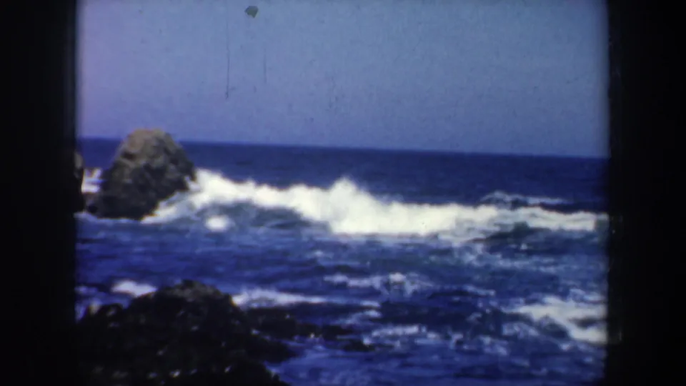 1957 San Francisco coastal frame with a large dark rock, white-capped surf, blue water, and clear sky on 8mm film.