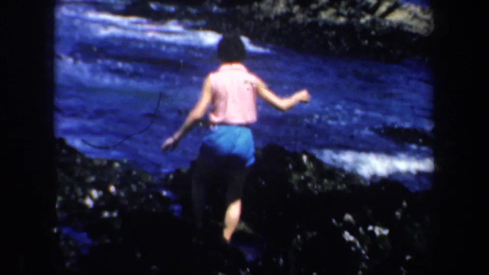 A woman in a pink top and blue shorts carefully crosses dark rocks near surf in 1957 San Francisco home-movie footage.
