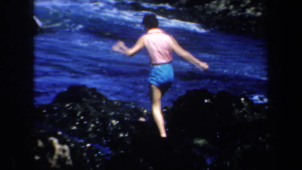 The woman continues across black coastal rocks, seen from behind against blue water and white surf in San Francisco archival footage.