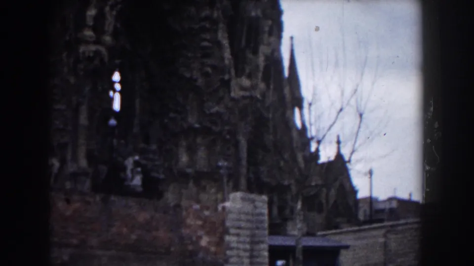 1959 Barcelona 8mm frame with shadowed church facade, low wall, bare tree branches, and a narrow view of nearby structures.