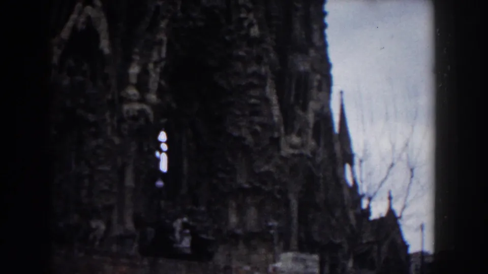 1959 Barcelona home-movie frame showing the church facade cropped close, with spire silhouettes, bare branches, and dark 8mm borders.